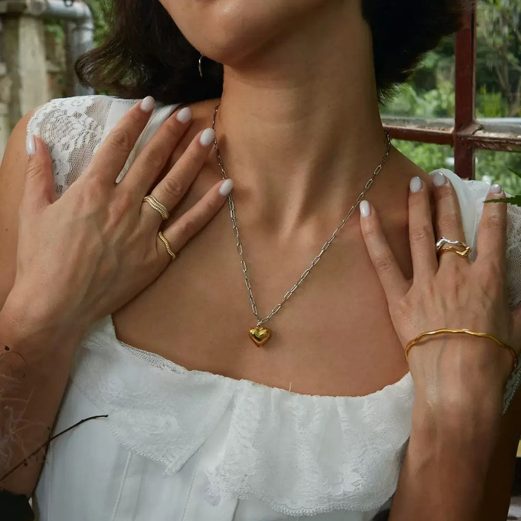 Woman wearing a gold necklace with a heart pendant, surrounded by greenery.