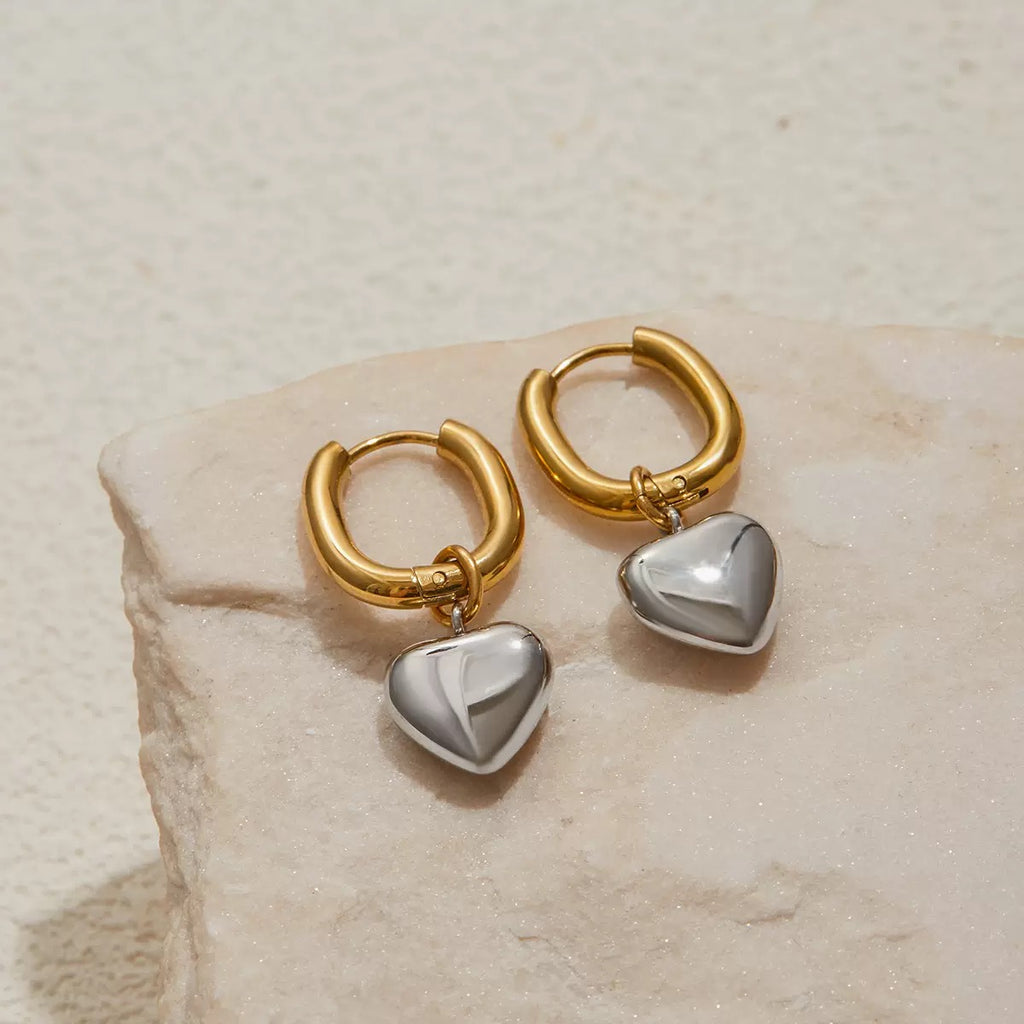 Gold hoop earrings with heart-shaped silver charms on a beige stone surface