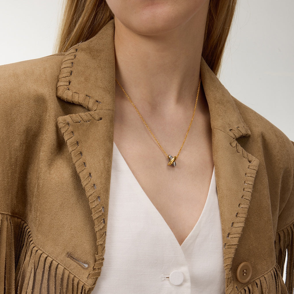 Person wearing a gold necklace with a heart pendant, brown suede jacket, and white blouse.