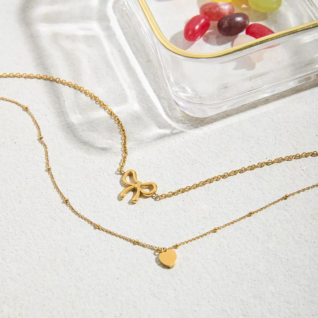 Two gold necklaces with bow and heart pendants on a white surface.