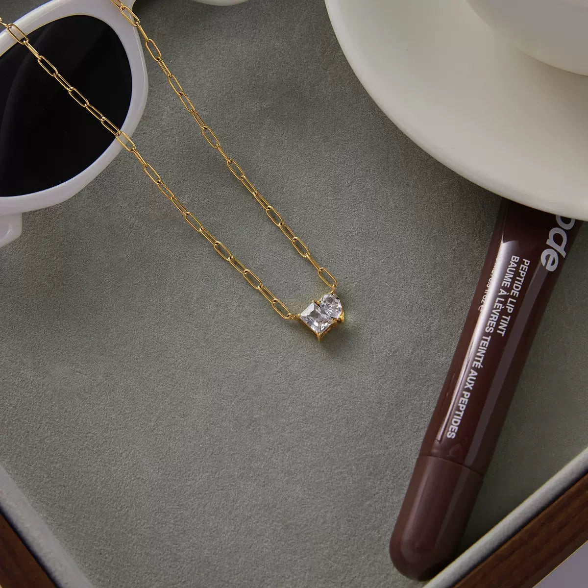 Gold necklace with a diamond pendant on a gray surface next to a lip balm and a white cup.