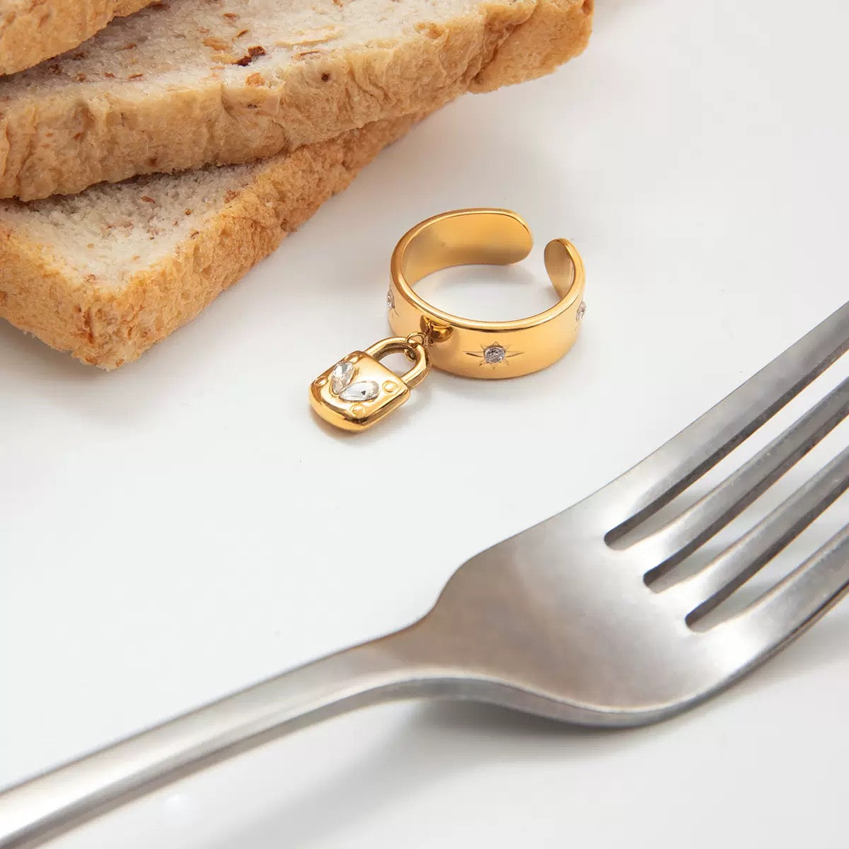 Gold ring with a lock charm on a white surface with bread and a fork.