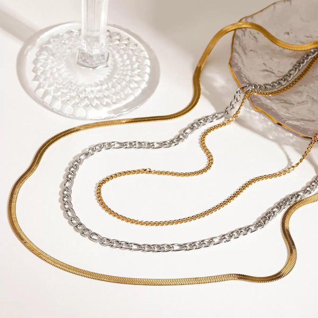 Gold and silver chain necklaces on a white surface with a glass of water in the background.