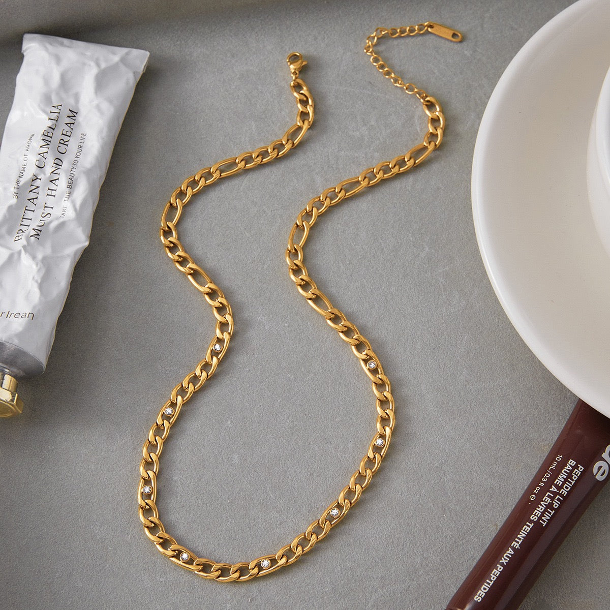 Gold chain on a gray surface with a tube of cream and a white cup.